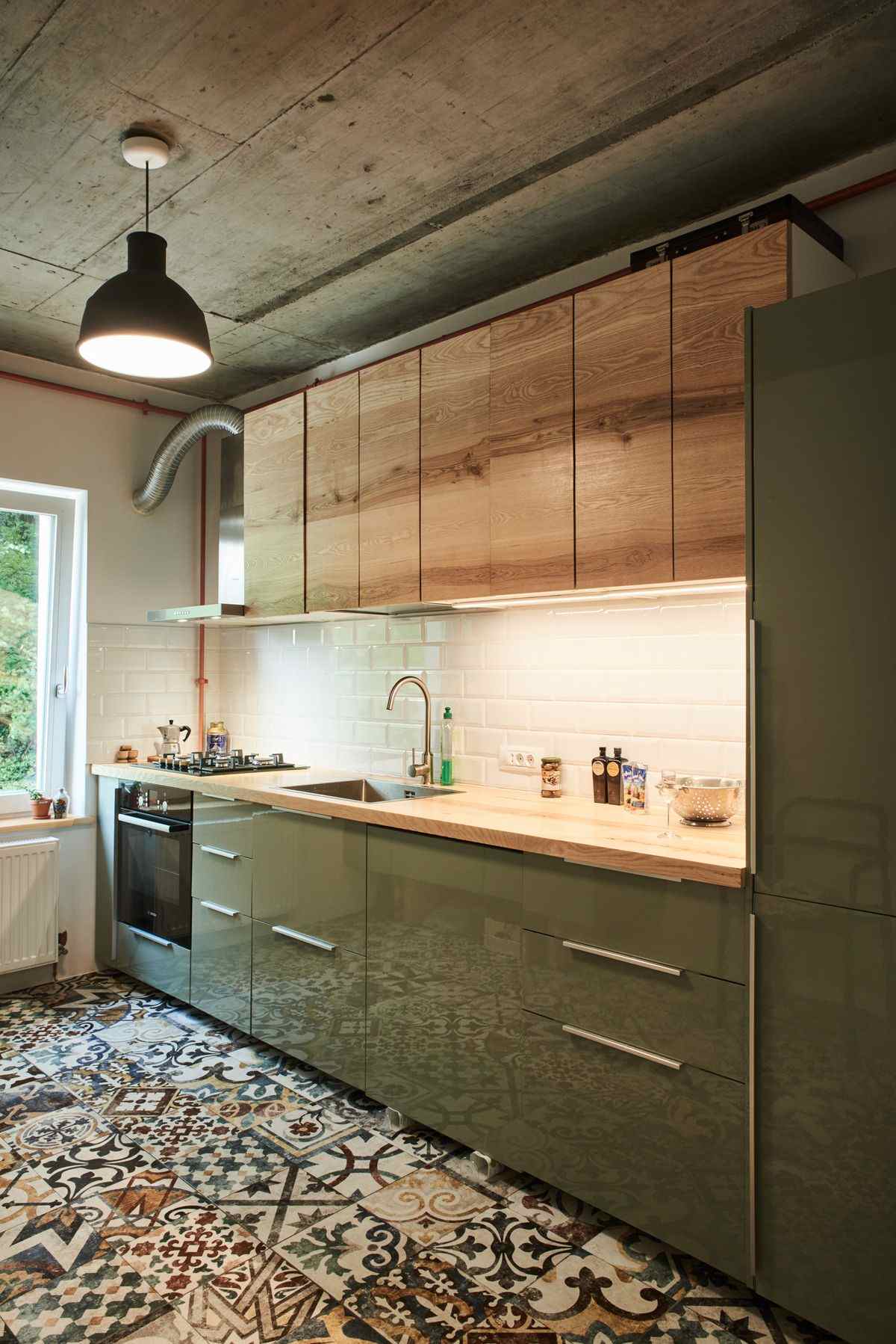 Vedere cu mobila de bucatarie cu fronturi verde oliv si corpuri superioare suspendate cu usi din lemn masiv de frasin. Pardoseala are motive florale geometrice, tavanul este din beton brut, cu o lampa moderna suspendata. In fundal este o fereastra ce ilumineaza incaperea.