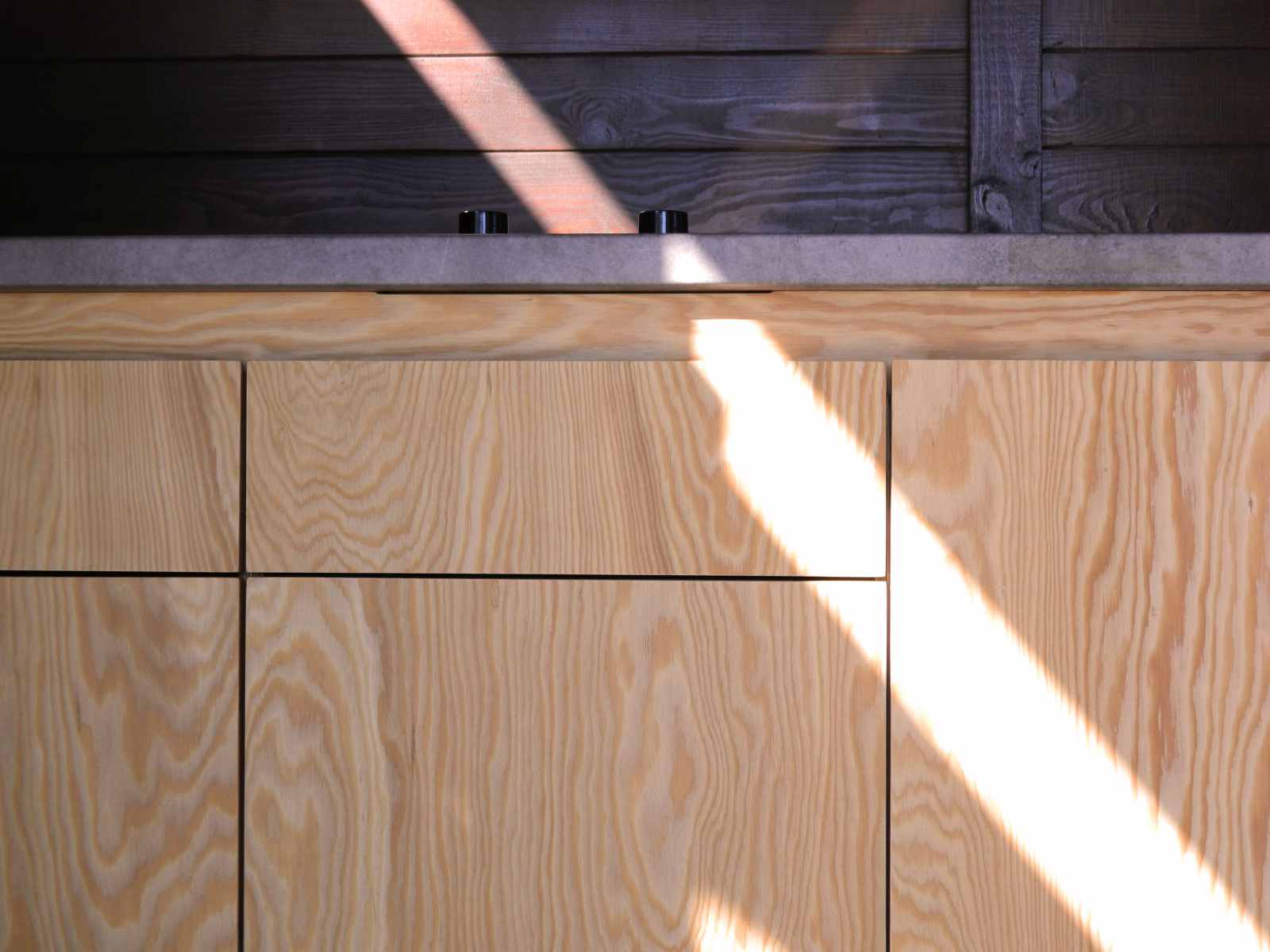 Fronturi de sertare si usi din placaj de larice. Pe blat se observa butoanele plitei. O raza de lumina cade oblic pe mobila de bucatarie