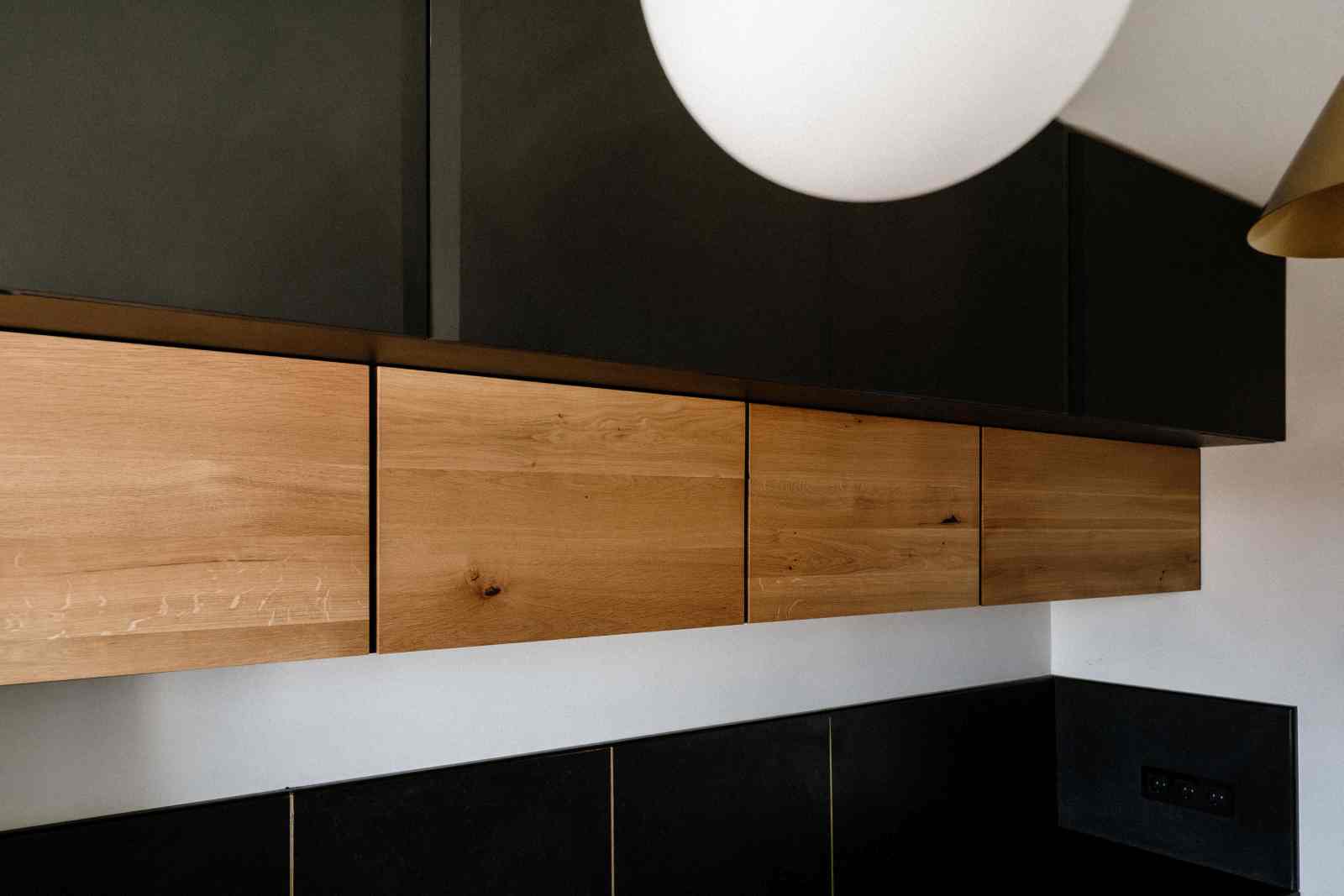 Corpuri de bucatarie suspendate pe doua niveluri prinse, cu usi din lemn de stejar si usi negre la partea superioara. In tavan este suspendata o lampa rotunda, iar blatul de bucatarie si contrablatul sunt de culoare inchisa cu insertii de alama.